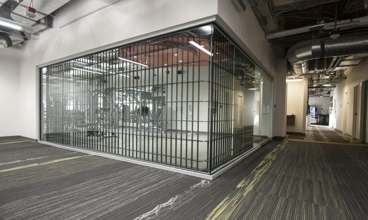 A modern glass-walled gym room with metal bars viewed from a hallway with exposed ceilings and patterned carpet.