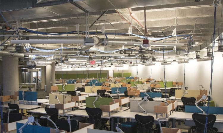 A large, empty open-plan office with rows of desks, colorful dividers, office chairs, and exposed ceiling pipes and wires.
