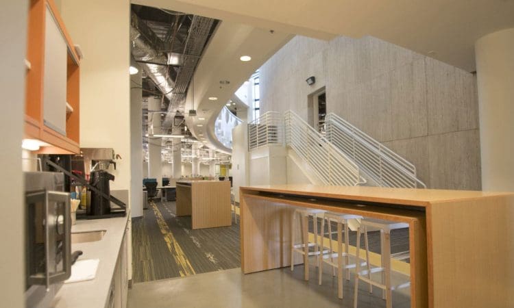 Modern office kitchen area with a long wooden table, white chairs, appliances on the counter, and a staircase in the background.