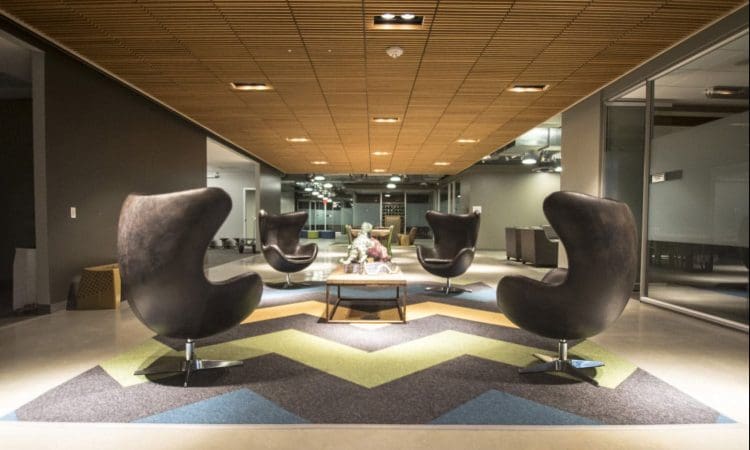 Modern office lounge with four black egg chairs around a coffee table on a chevron-patterned rug, under a wooden panel ceiling with glass walls in the background.