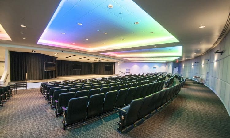 Empty modern auditorium with rows of black seats facing a stage, two large screens, and a ceiling lit with multicolored lights.