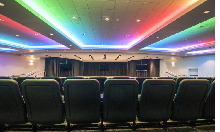 Rows of empty auditorium seats face a stage with monitors, under a ceiling lit by colorful LED lights in green, blue, and red hues.