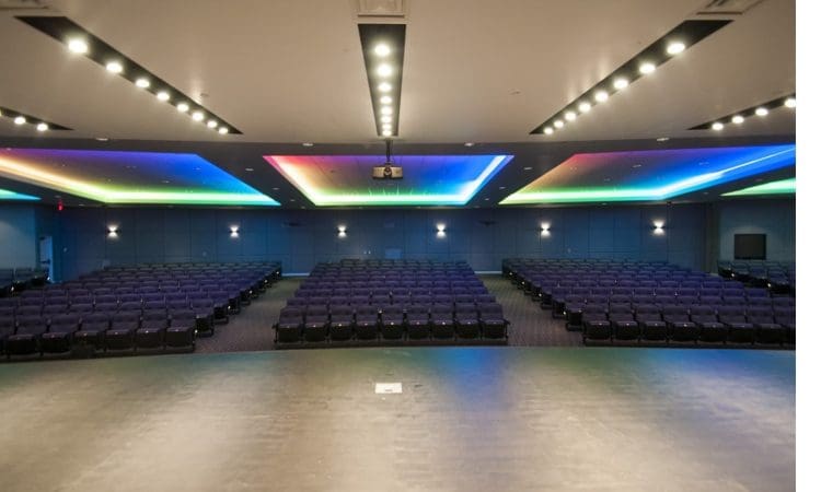 View from stage of an empty auditorium with rows of purple seats and a ceiling lit by colored LED lights.